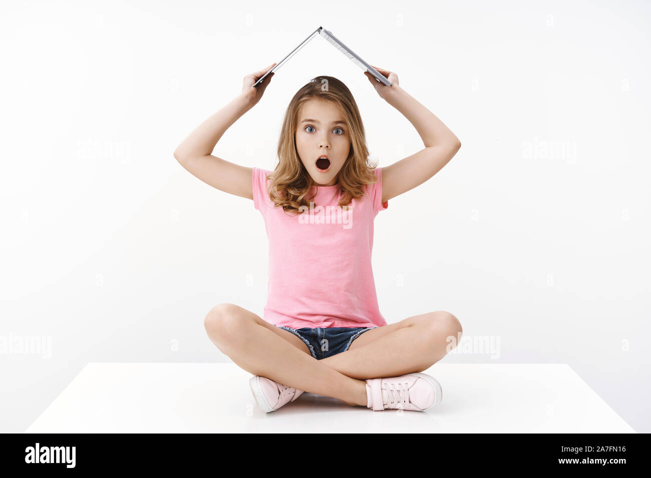 Amused and shocked blond young girl raise laptop under head, use computer like roof, open mouth gasping amazed and impressed, sit on floor with Stock Photo
