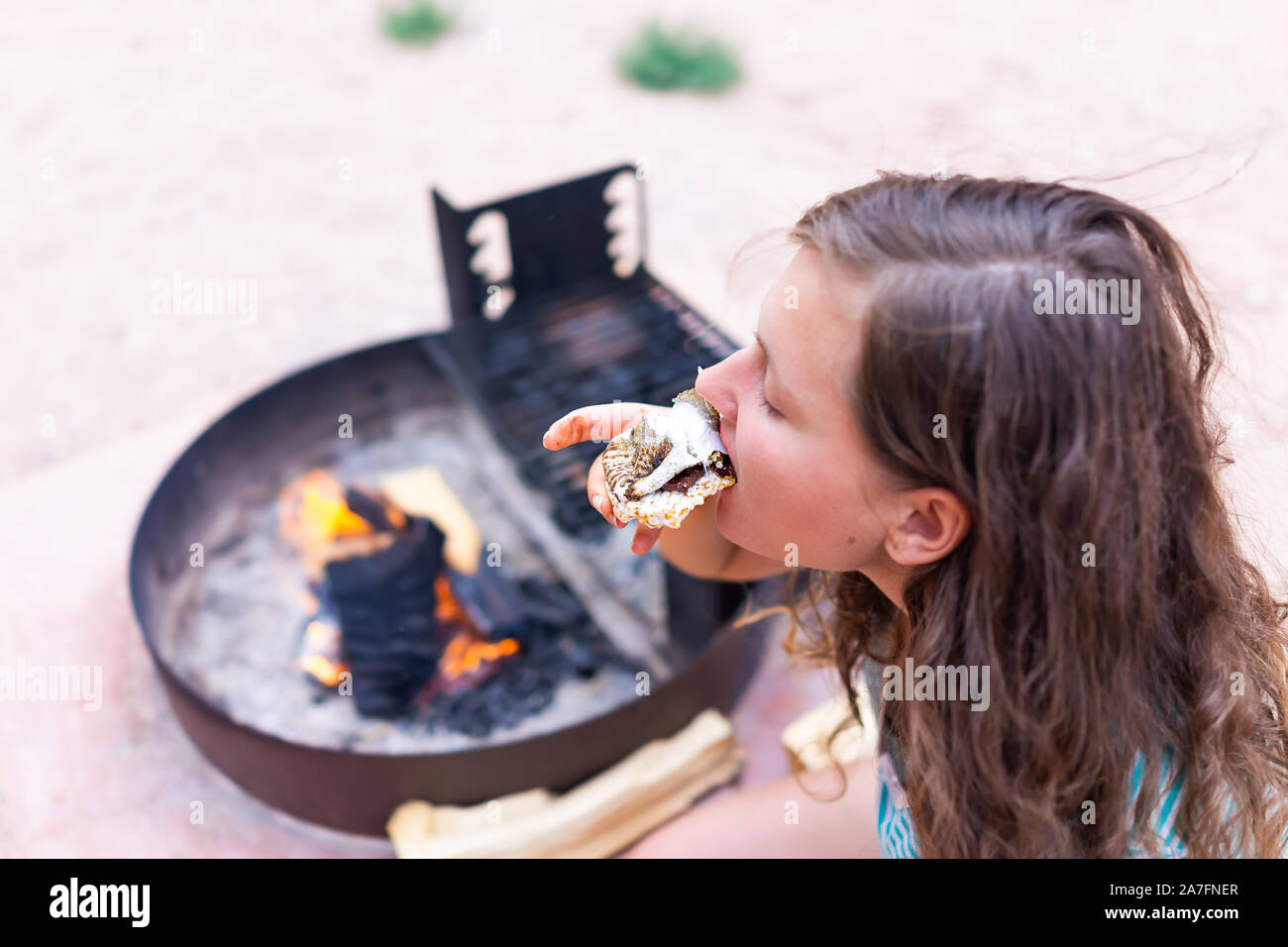 Young woman eating gooey roasted marshmallows smores with chocolate and rice cake cracker by fire in campground campfire grill Stock Photo