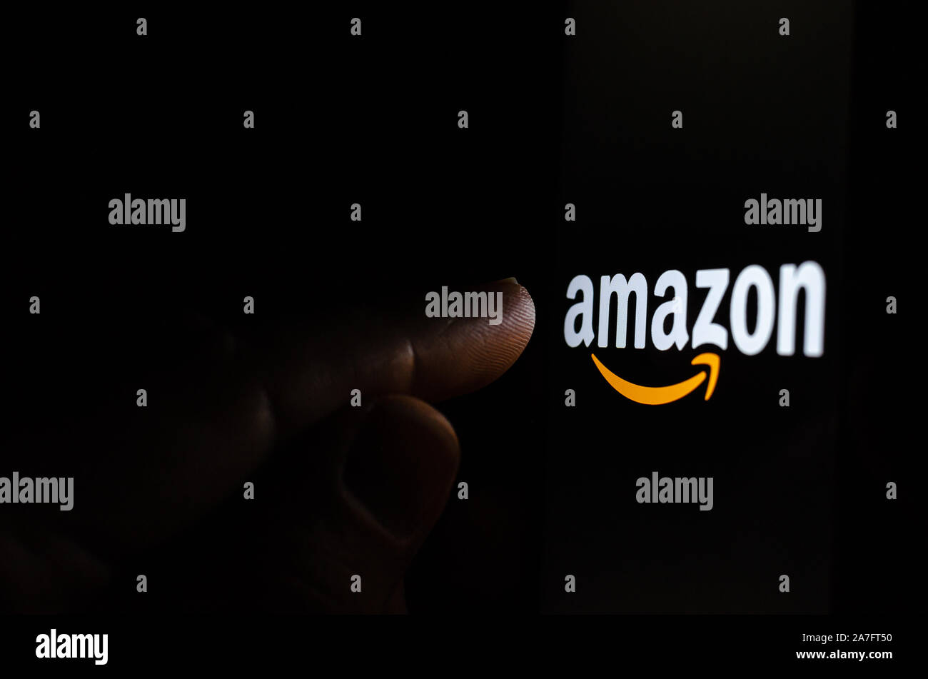 Amazon logo on a smartphone screen in a dark room and a finger pointing at it. Stock Photo