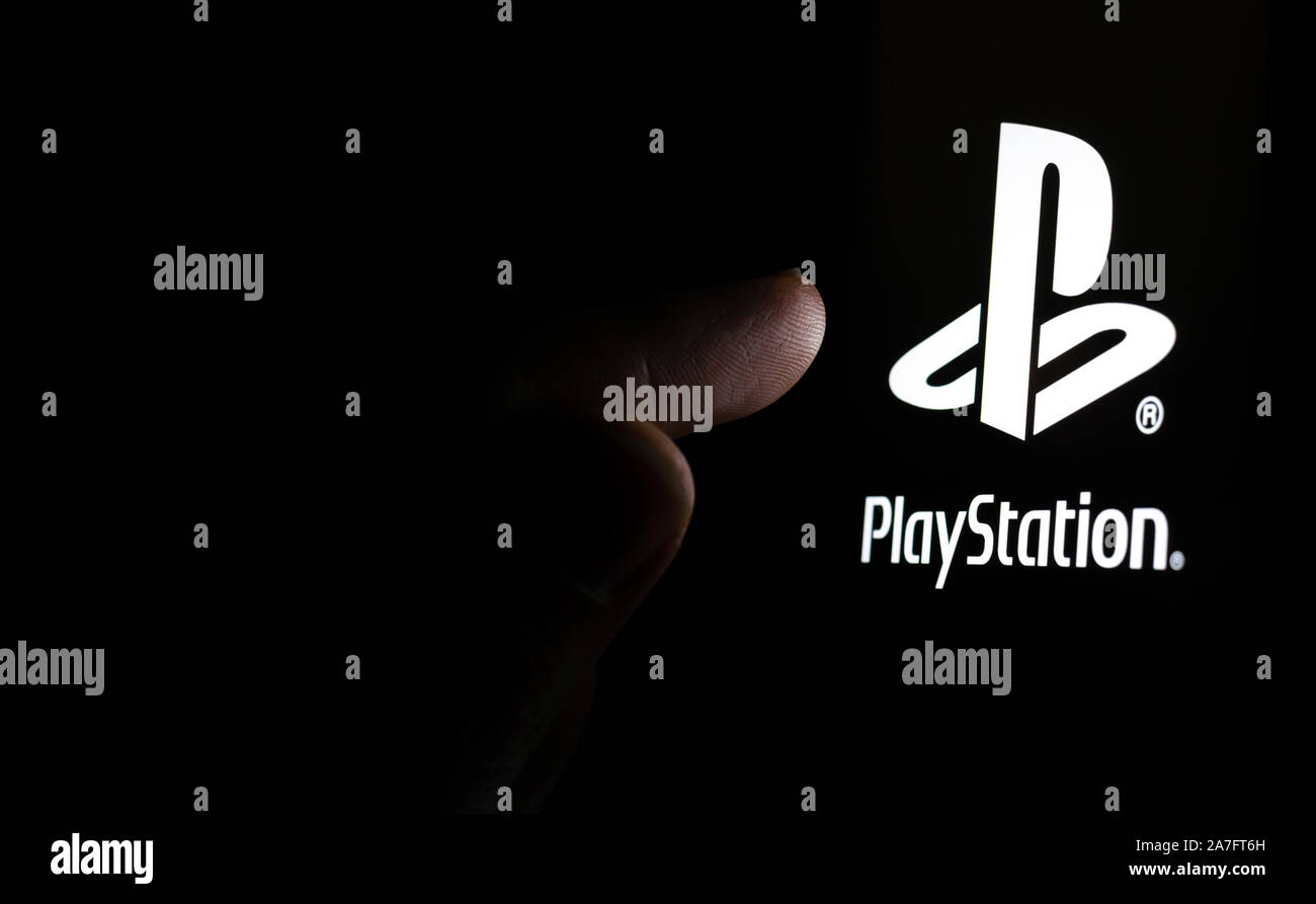 PlayStation logo on a smartphone screen in a dark room and a finger touching it. Stock Photo