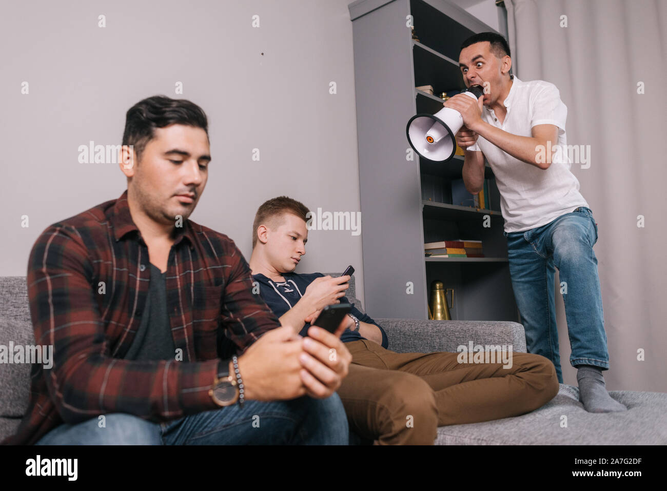 Young man shouting through a megaphone at friends which using a mobile phone. Stock Photo
