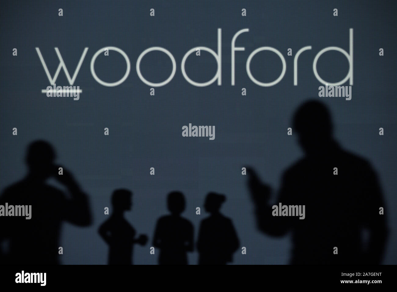 The Woodford Patient Capital Trust logo is seen on an LED screen in the background while a silhouetted person uses a smartphone (Editorial use only) Stock Photo