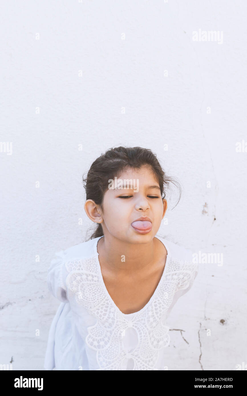 Little funny girl in white dress shows off her tongue. Cute kid girl sticking tongue out. Portrait of angry and unhappy young lady open mouth and prot Stock Photo