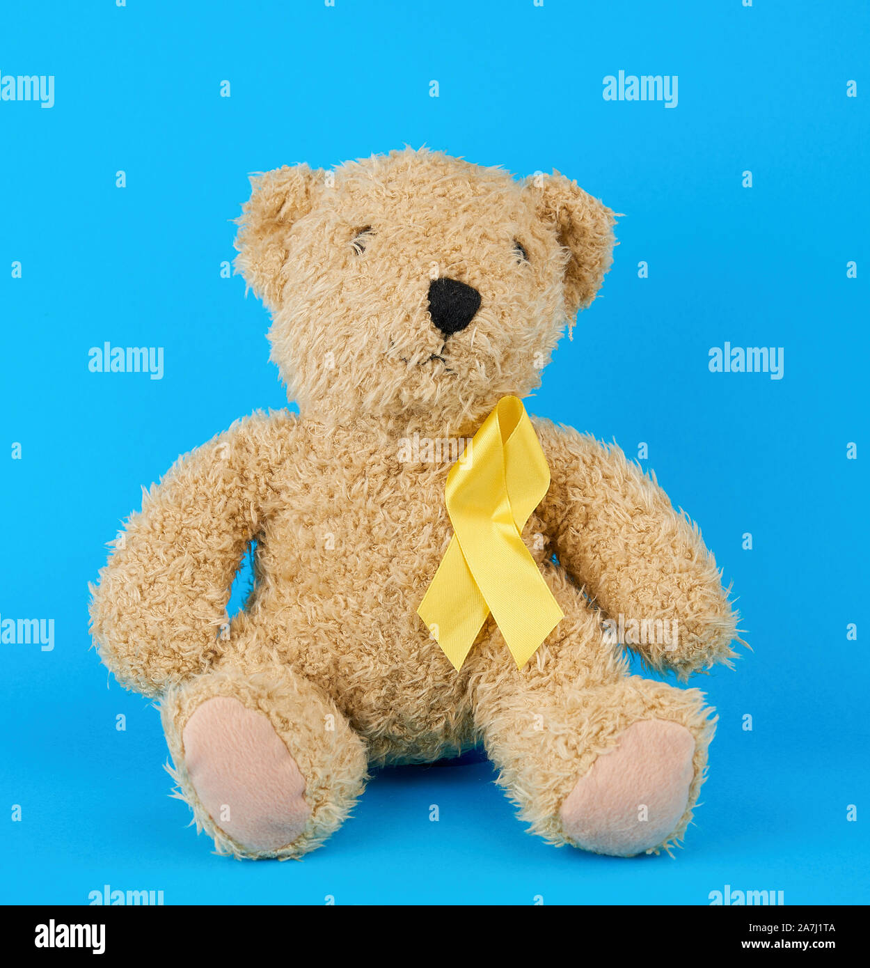 brown teddy bear sits and holds in his paw a yellow silk ribbon on a blue background, concept of the fight against childhood cancer. problem of suicid Stock Photo