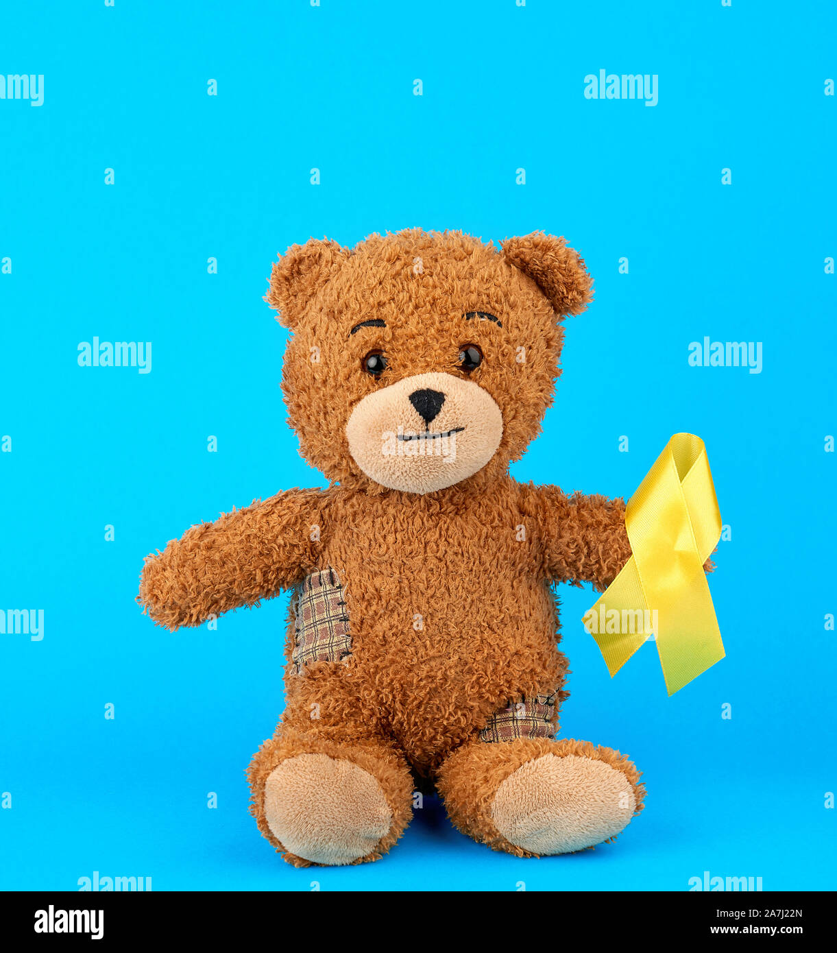 brown teddy bear sits and holds in his paw a yellow silk ribbon on a blue background, concept of the fight against childhood cancer. problem of suicid Stock Photo