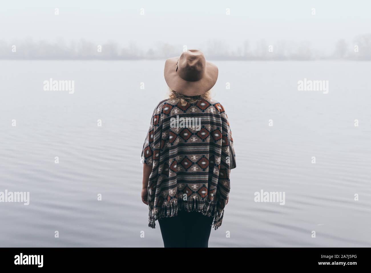 Woman in hat and poncho standing on the river bank and looking towards autumn forest covered with morning fog. Stock Photo