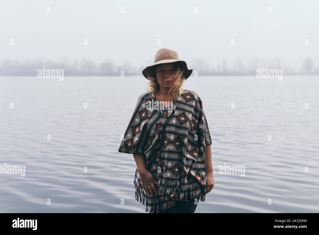 Woman in hat and poncho standing on the river bank and looking towards autumn foggy forest. Stock Photo