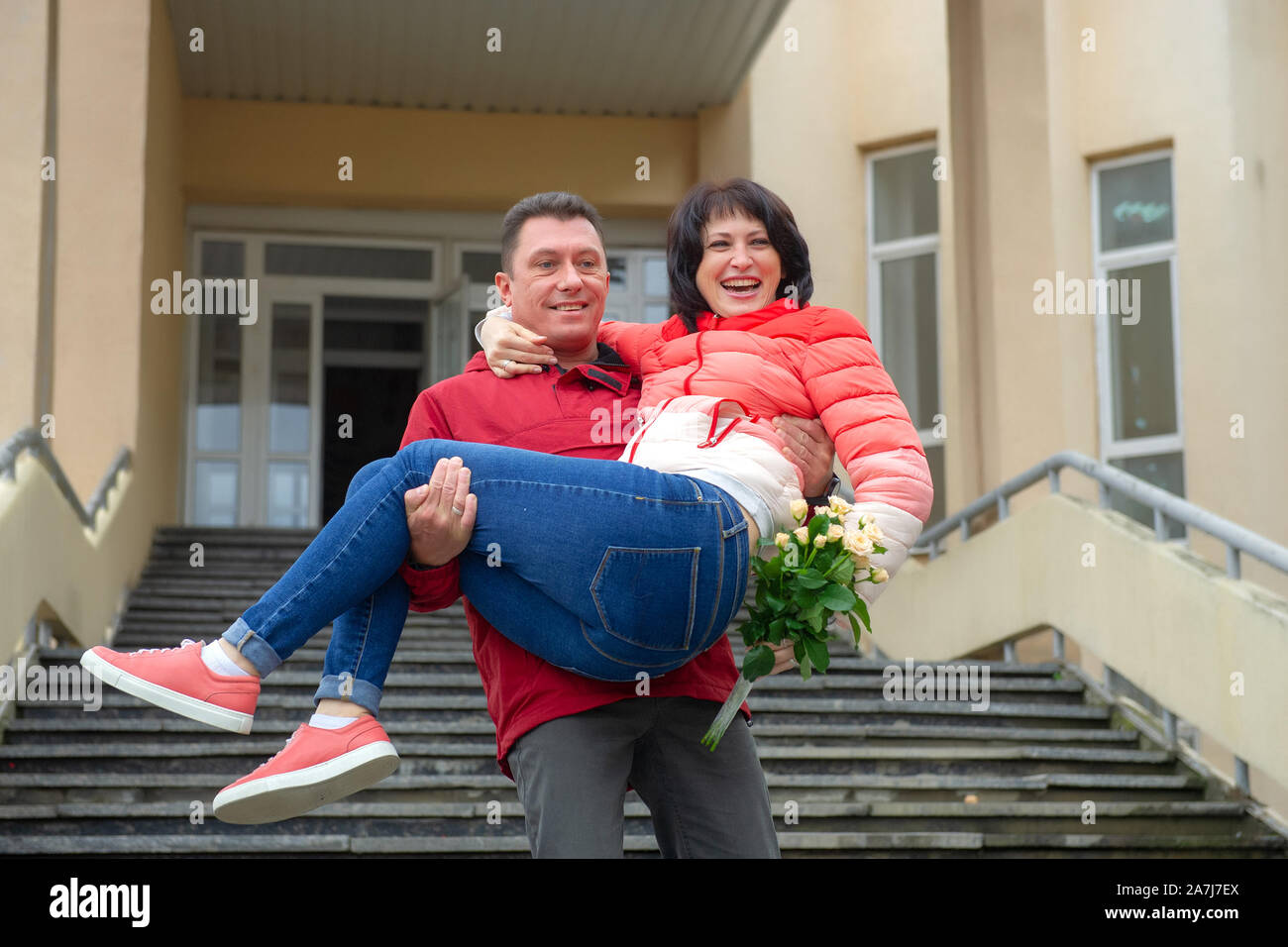 A man holds a woman in her arms with a bouquet of flowers. Woman in the arms of a man with a bouquet of flowers. Stock Photo