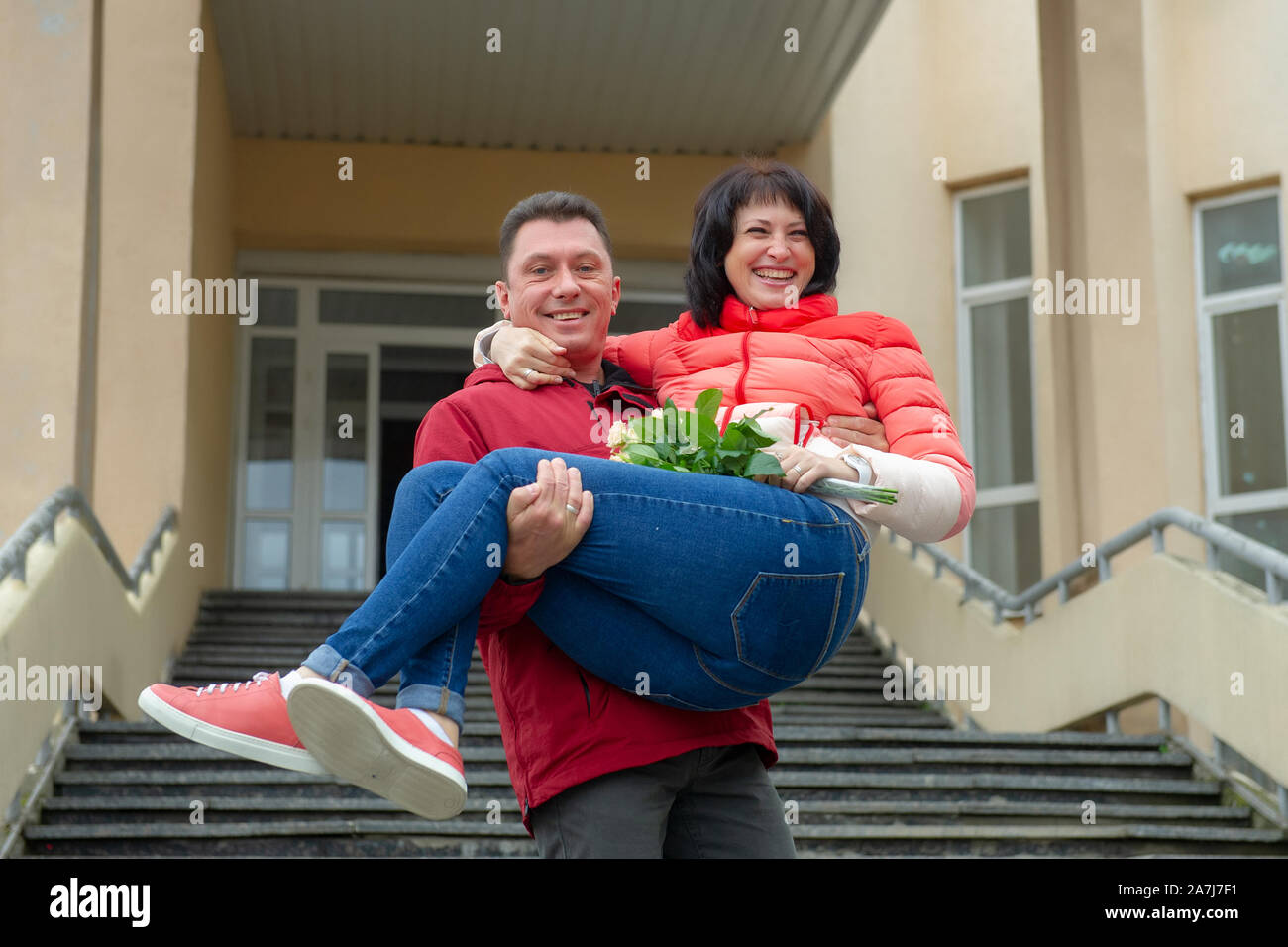 A man holds a woman in her arms with a bouquet of flowers. Woman in the arms of a man with a bouquet of flowers. Stock Photo