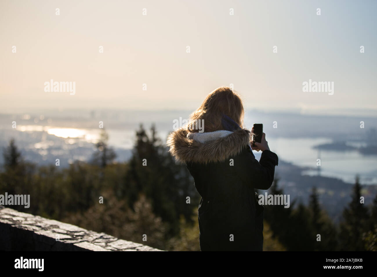 A young woman in a winter coat at the Cypress Mountain lookout taking a photo of downtown Vancouver on her cell phone. Stock Photo