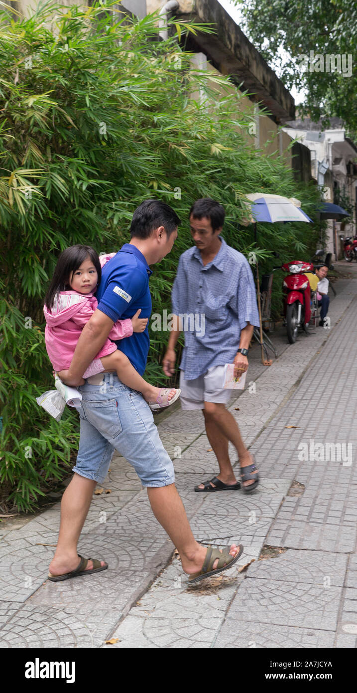 Father carrying little girl on back while walking down Saigon tree lined sidewalk on summer day. Stock Photo