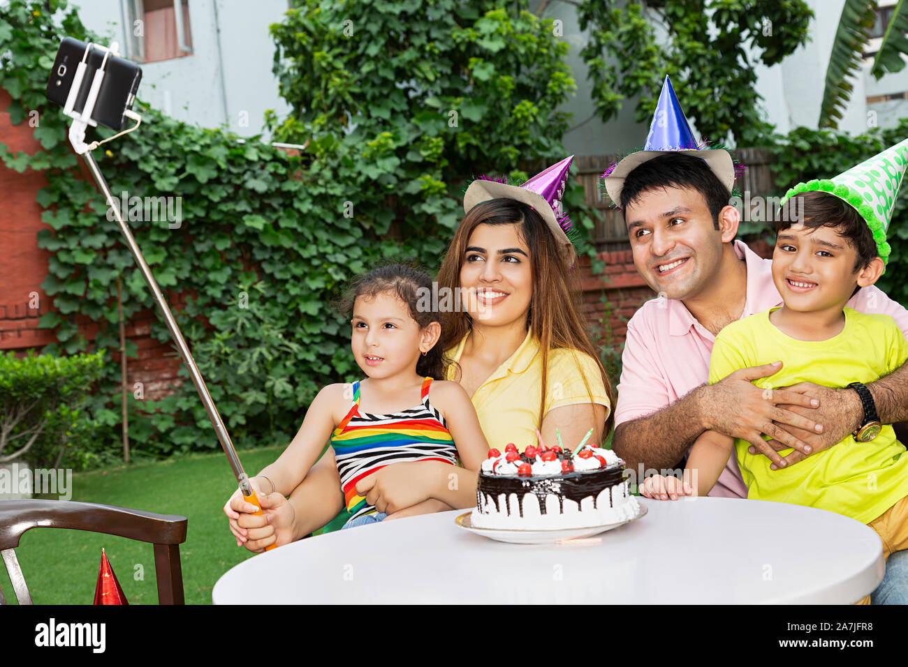 Happy-family Parents And Two-Children taking Selfie picture with smartphone on selfie stick during birthday celebration in-Courtyard Of-Their Home Stock Photo