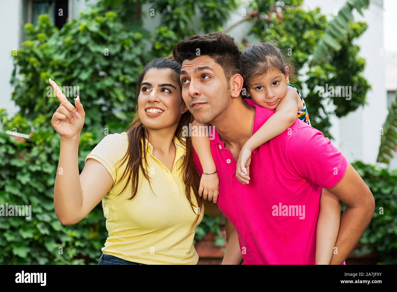Happy Indian Family Father carrying his Little daughter on-piggyback With Mother Pointing Finger Somewhere in-Outdoor At-park Stock Photo