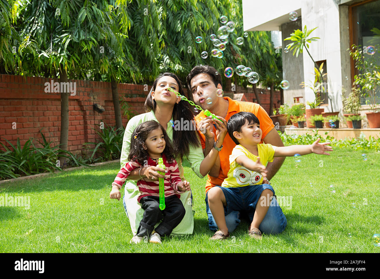 Happy Young Family Parents And Two Kids blowing into bubble wand making soap balloons in-courtyard near their houseÂ Stock Photo