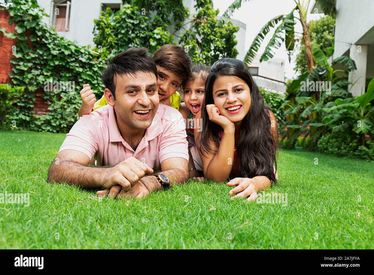 Happy-family Father Mother And Two-Kids Lying-On-Grass Relaxing Together Having Fun Enjoying in-garden Of-their House Stock Photo