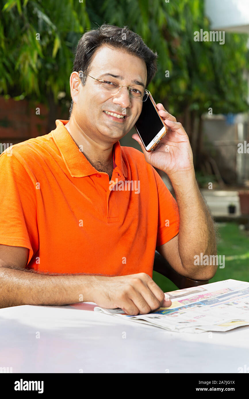Portrait Of Mid-Adult Man Reading Newspaper While Talking On Smart Phone in Outdoor garden Stock Photo