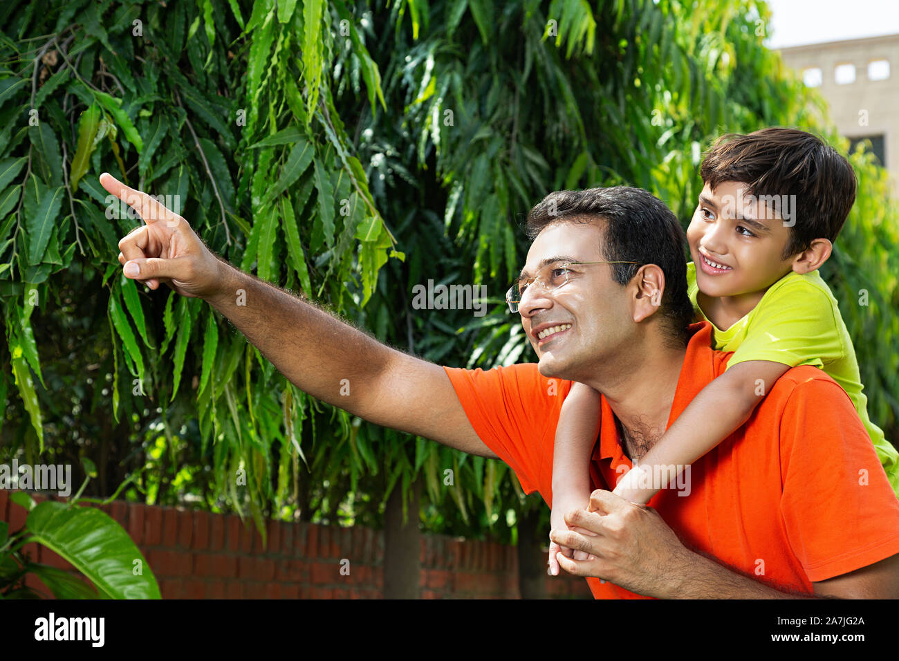 Happy Indian father holds his Kid son piggyback ride on his shoulders points at something in-Outdoor garden Stock Photo