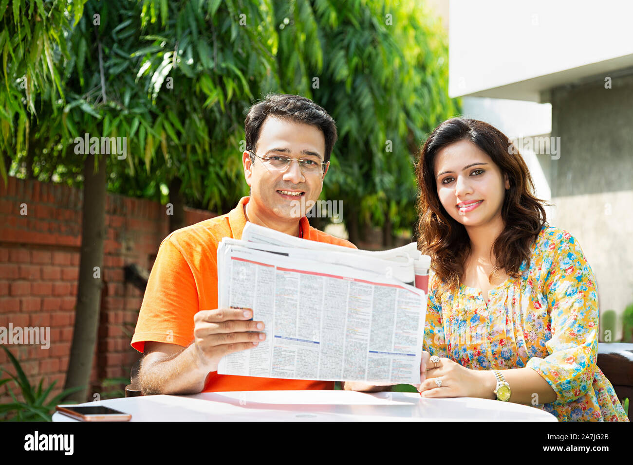 Happy Indian Married Couple Sitting-on-Table Reading Newspaper Together in-Courtyard of-their home in-Morning Stock Photo