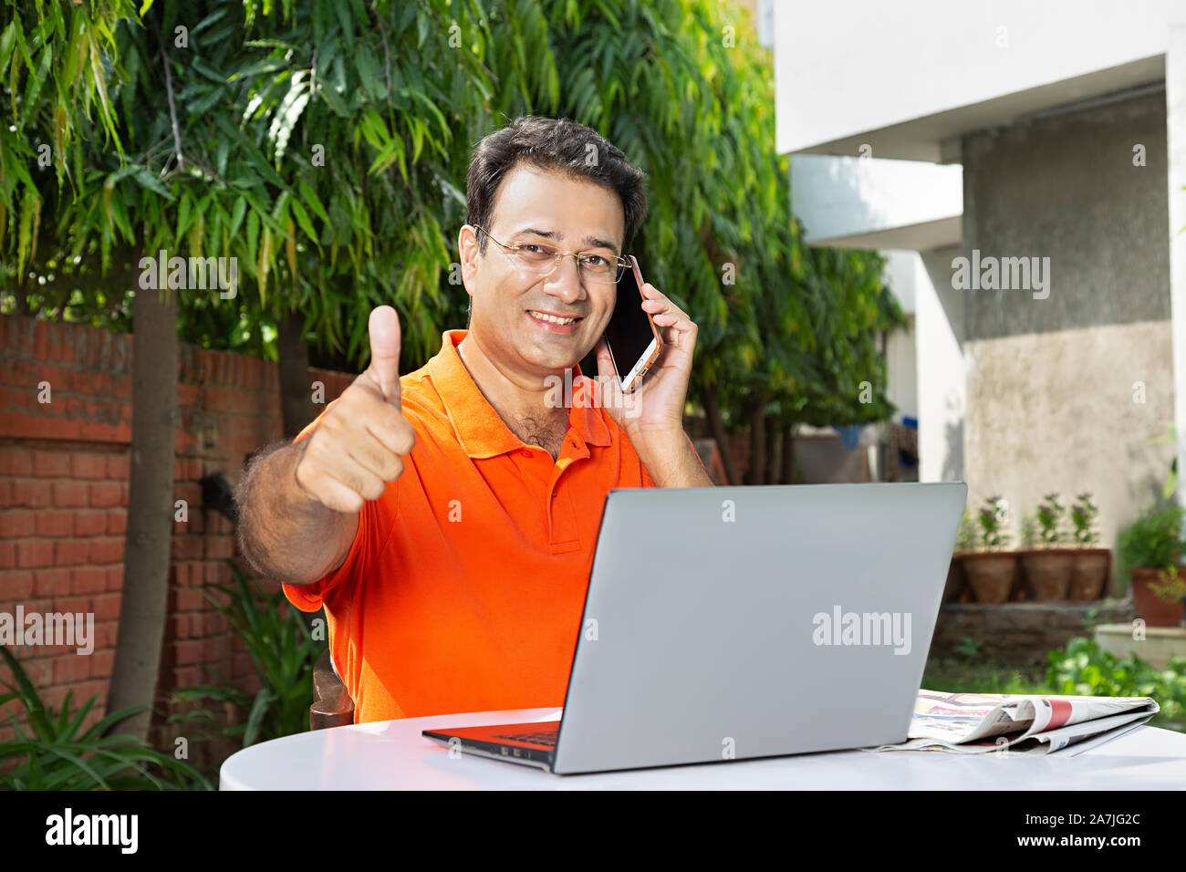 Happy Mid-Adult male Talking Mobile Phone And Showing Thumbs-up With laptop in-courtyard Near their house Stock Photo