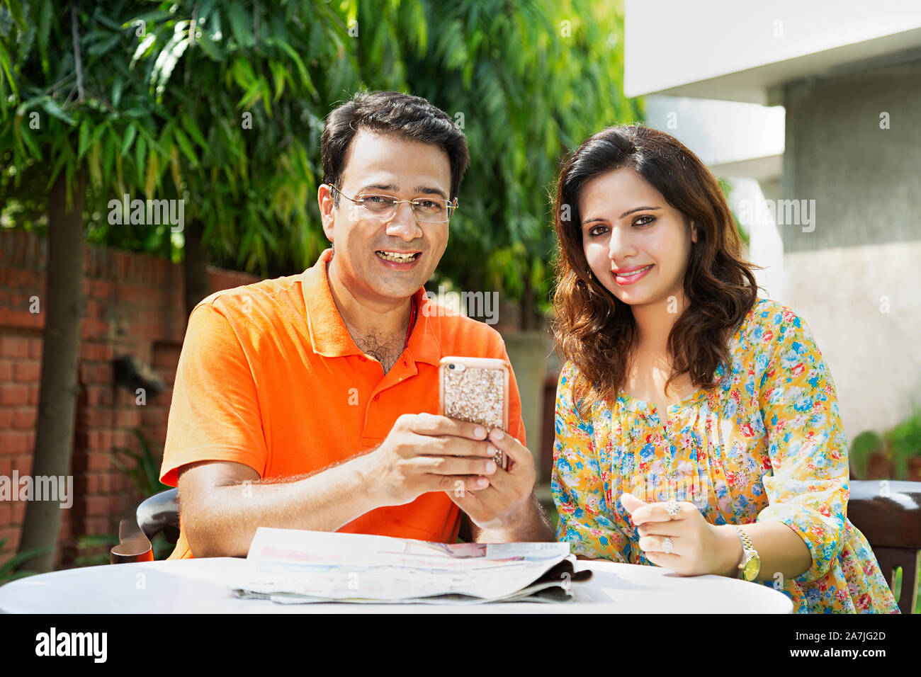 Portrait of Smiling Mid-adult Couple Sitting-on-table And using Mobile Phone in-courtyard of-their House Stock Photo