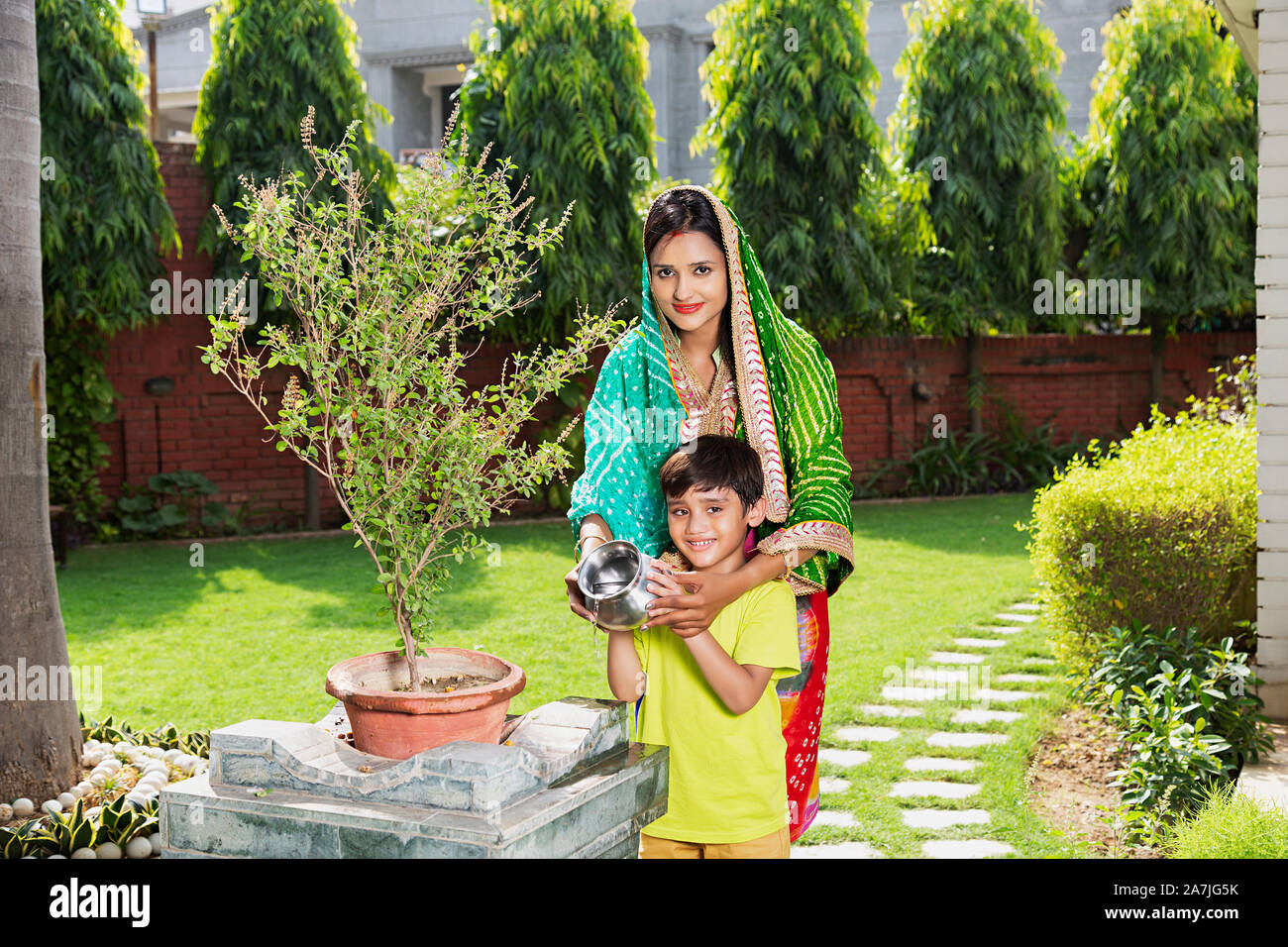 Indian Hindu Mother And Kid Son water-pouring in-holy Tulsi-Plant performing puja in-Courtyard Of-Their Home Stock Photo
