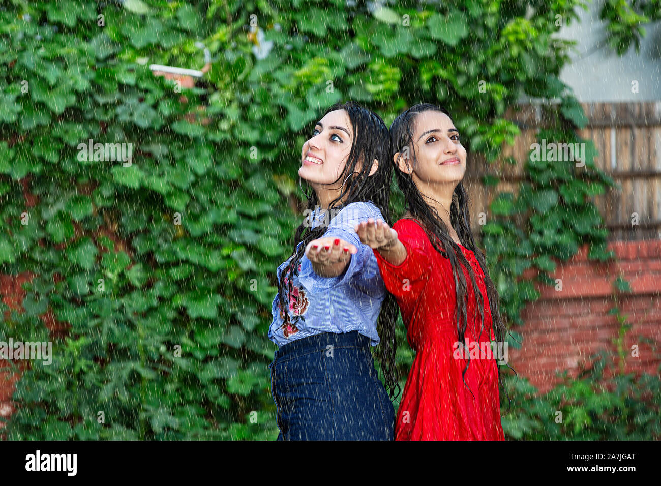 Happy Two Young Female Friends Wiyh Arms open Standing back-to-Back Enjoying the-rain in-garden Rainy weather Stock Photo