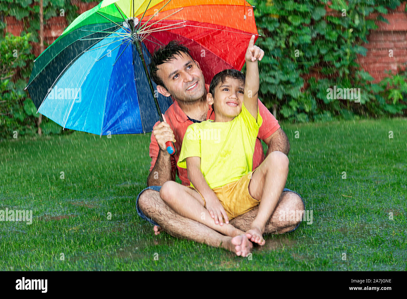 Happy-family Father And Kid boy Sitting-on-Grass With umbrella Pointing at Something in-The Rain in-Outdoor garden Stock Photo