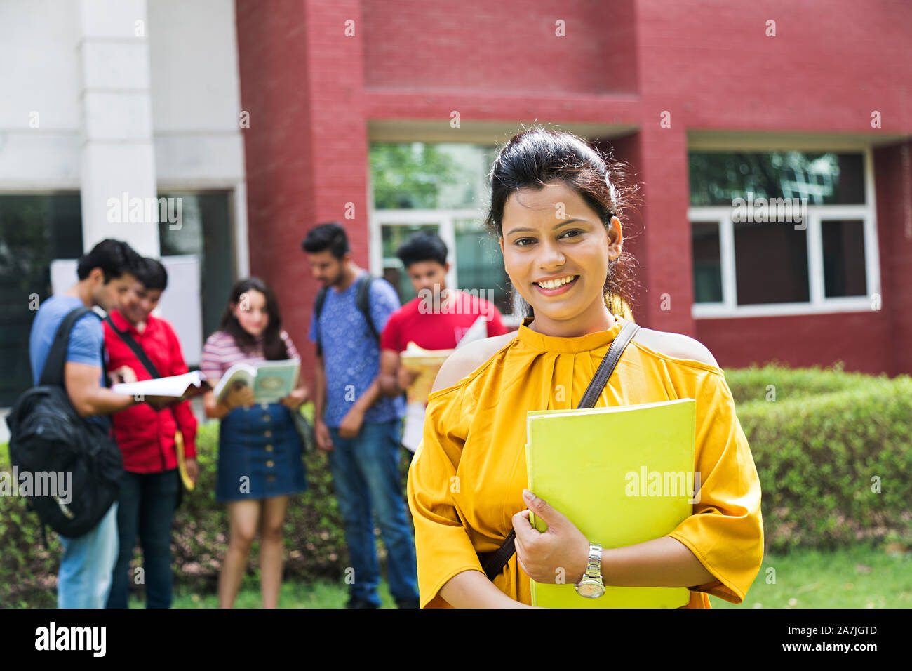 Happy Young Female College student Holding Book While Classmates In-The-background Outside Campus Building Stock Photo