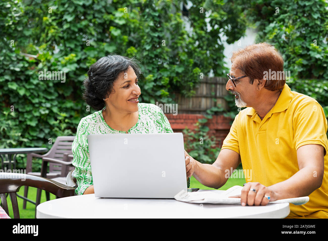 Happy indian Senior Couple Male And Female Using Laptop And With Newspaper in the courtyard of their house On Summer Morning Stock Photo
