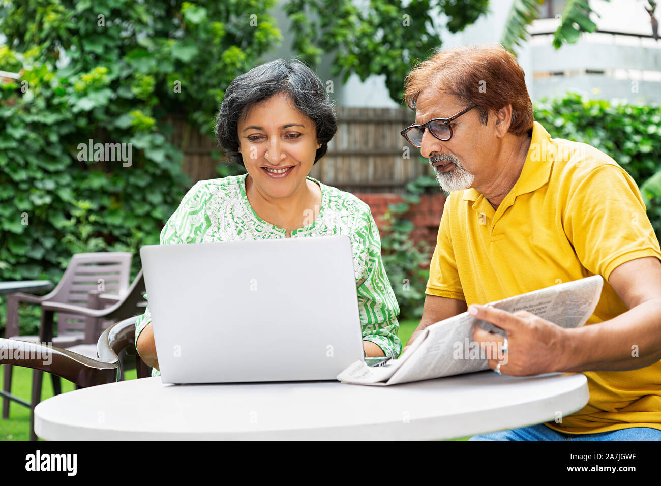 Indian Elderly Couple Male And Female Looking-at Laptop Screen With Newspaper Sitting In the courtyard Of Their home Stock Photo