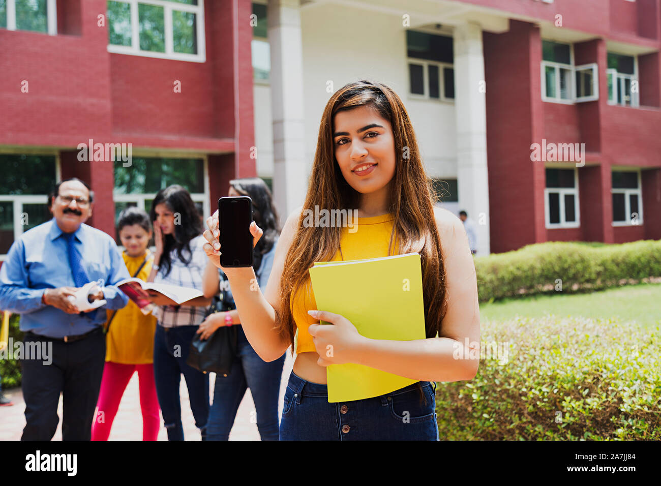 Young Female College Student Showing Cell-Phone E-Learning With Students And Teacher In-Background At-Campus Stock Photo