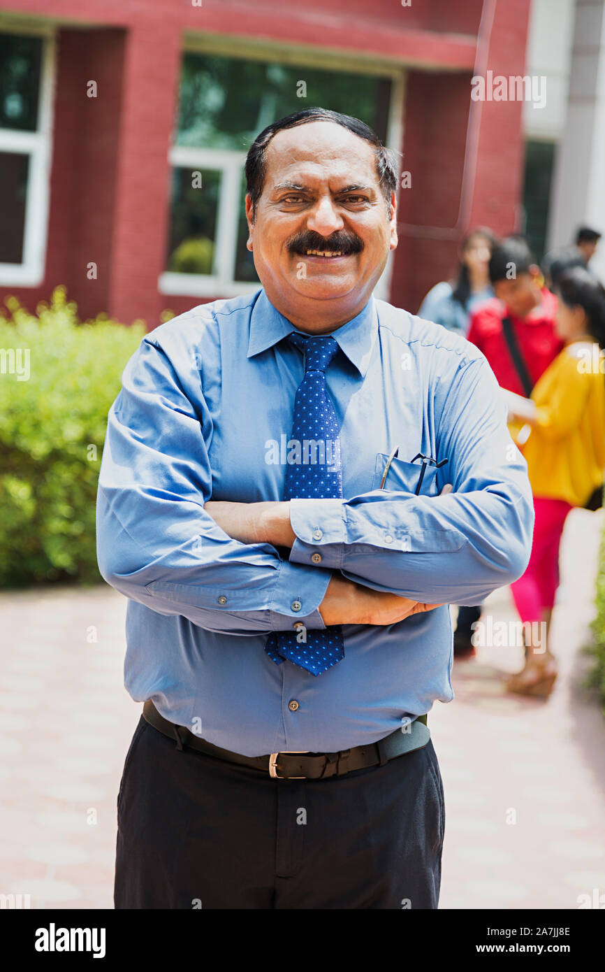 Senior male College Teacher With Arms-Folded Standing With People-In-The-Background In-Outside Campus Stock Photo