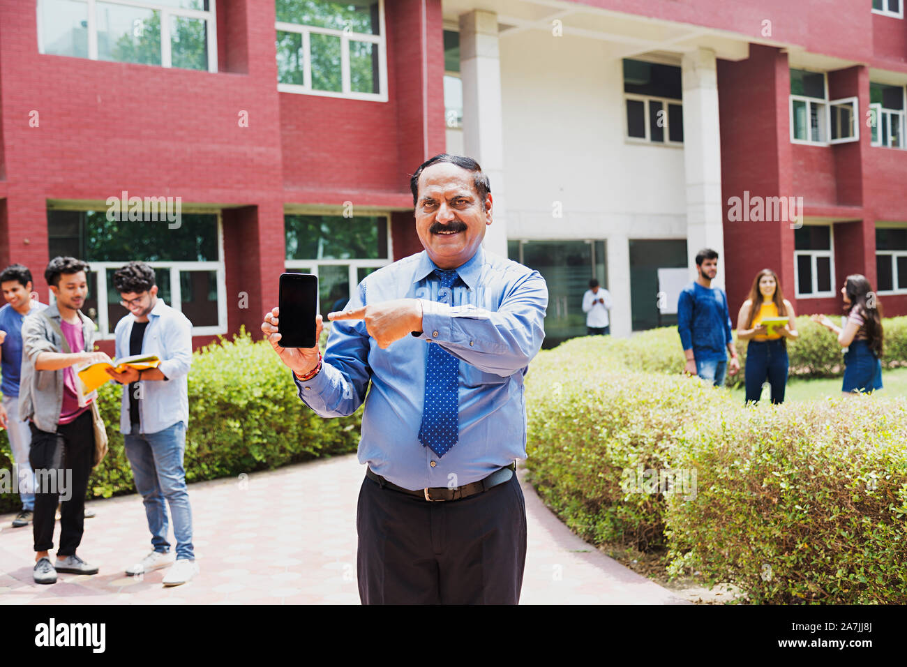 Senior man College Teacher Pointing Finger Showing Mobile-Phone With Students In-The-Background In-Outside Campus Building Stock Photo