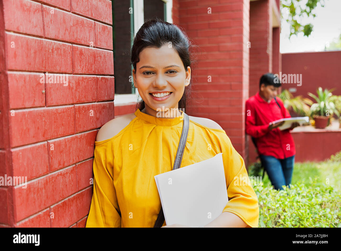 Indian Young Girl College Student holding Book Standing leaning against Wall College Campus Stock Photo