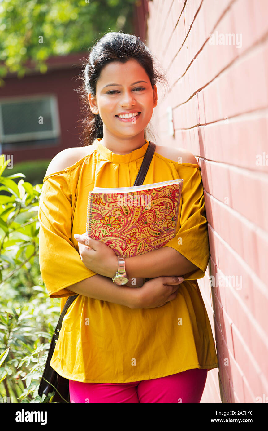 Young Female College Student holding Book Standing leaning against wall College Campus Building Stock Photo