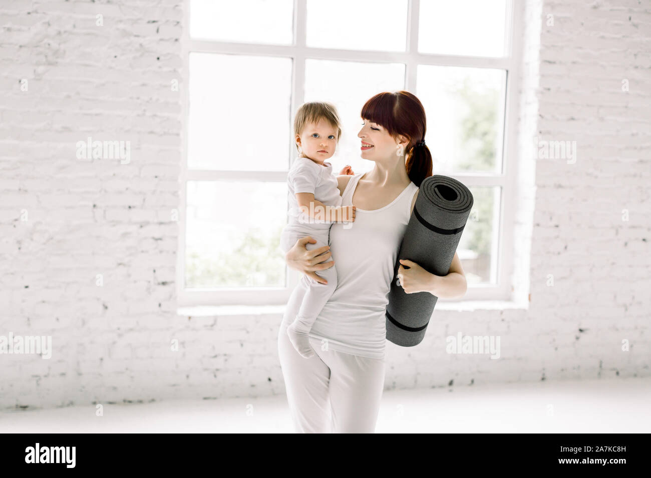 Sportive pretty young mother with little baby girl in white sport wear and yoga mat in hands, standing in light gym hall. Fitness, happy maternity and Stock Photo