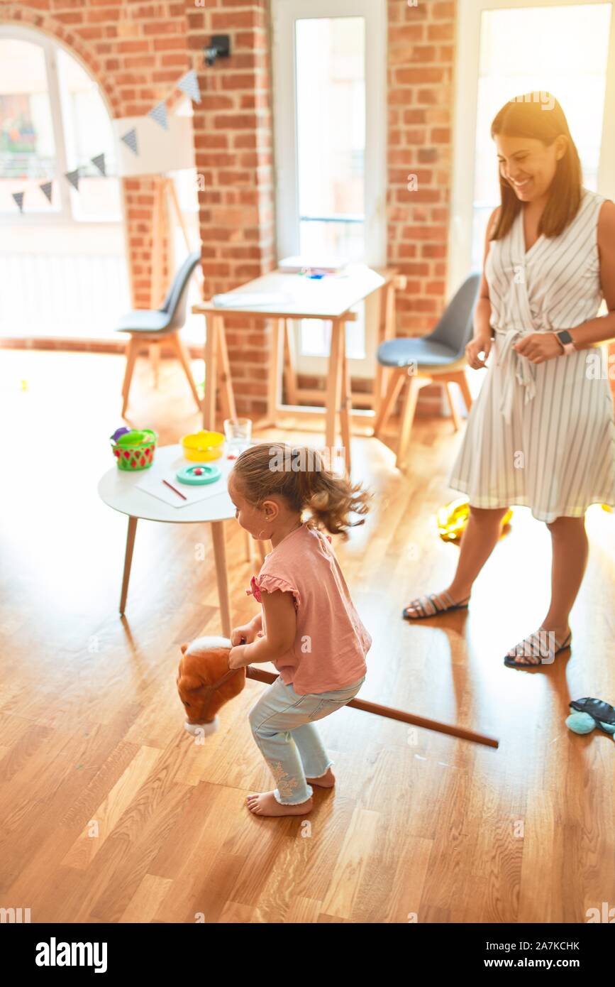 Beautiful teacher and blond student toddler girl ridding horse toy with stick at kindergarten Stock Photo