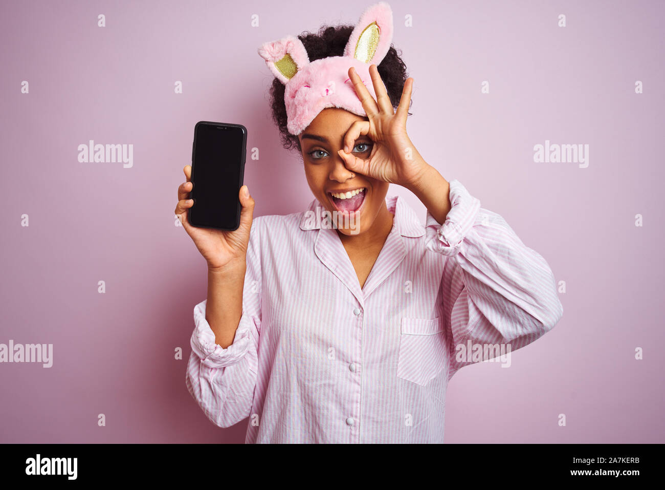 Afro woman wearing pajama and mask showing smartphone over isolated ...