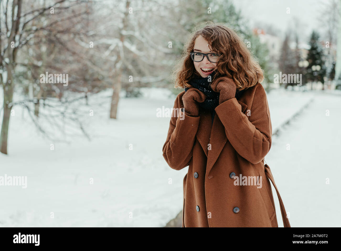 Young beautiful hipster woman in knitted scarf standing in the park and smiling. Winter fashion. Snow Stock Photo