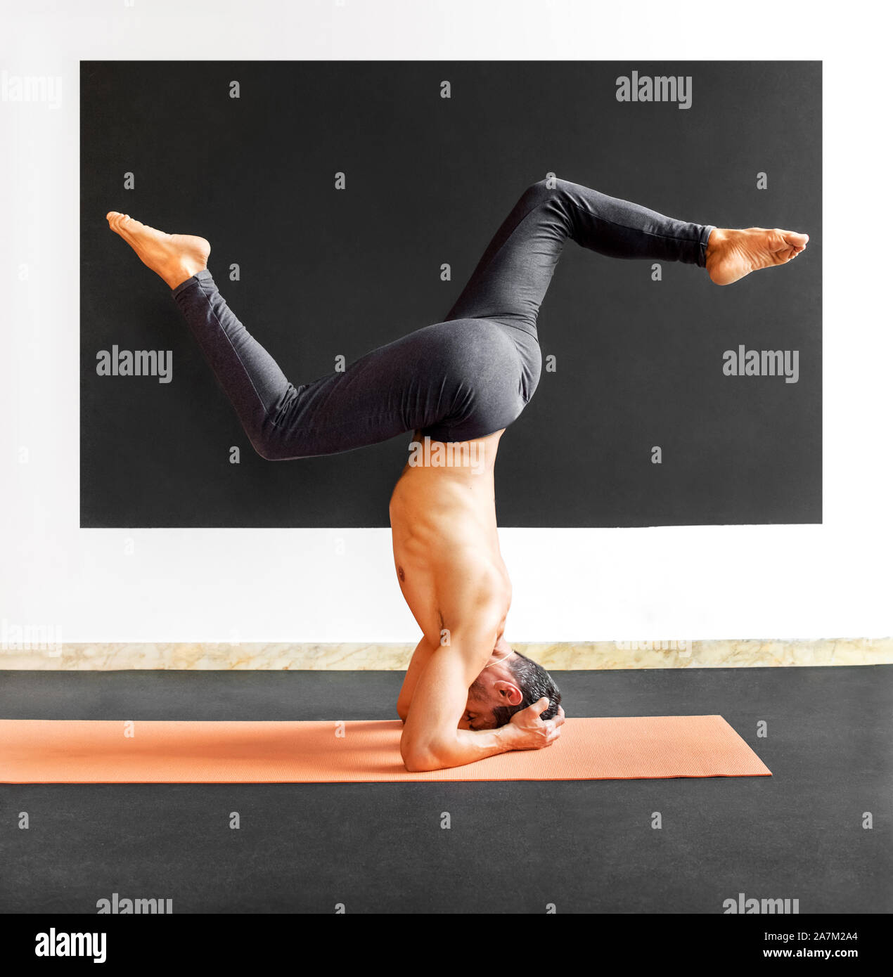 Man doing a sirsasana variation yoga headstand in a gym to align his body, improve blood flow and strengthen his muscles Stock Photo