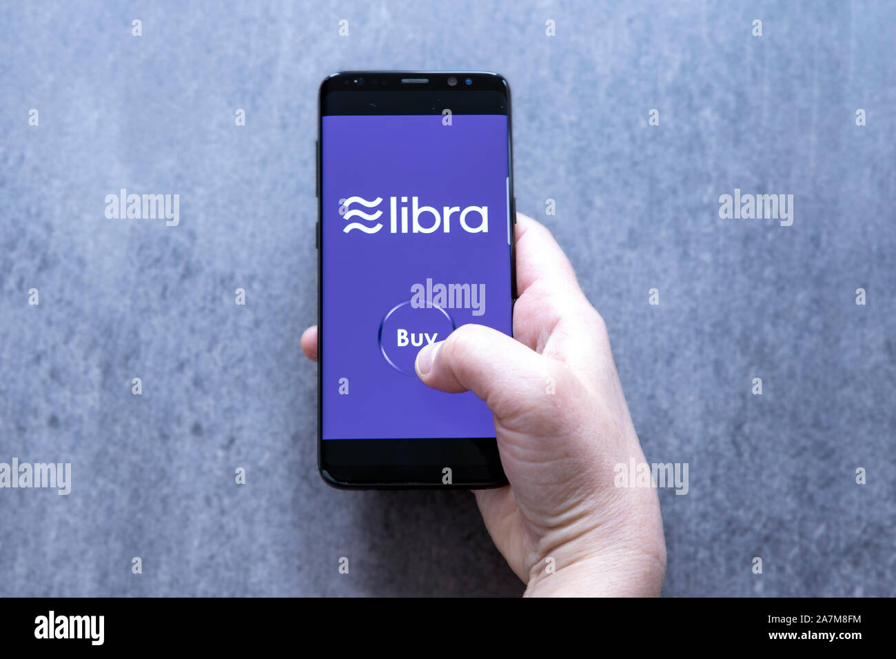 Hand holding smartfone with crypto currency logo Libra and buy button against gray blurred background. Stock Photo