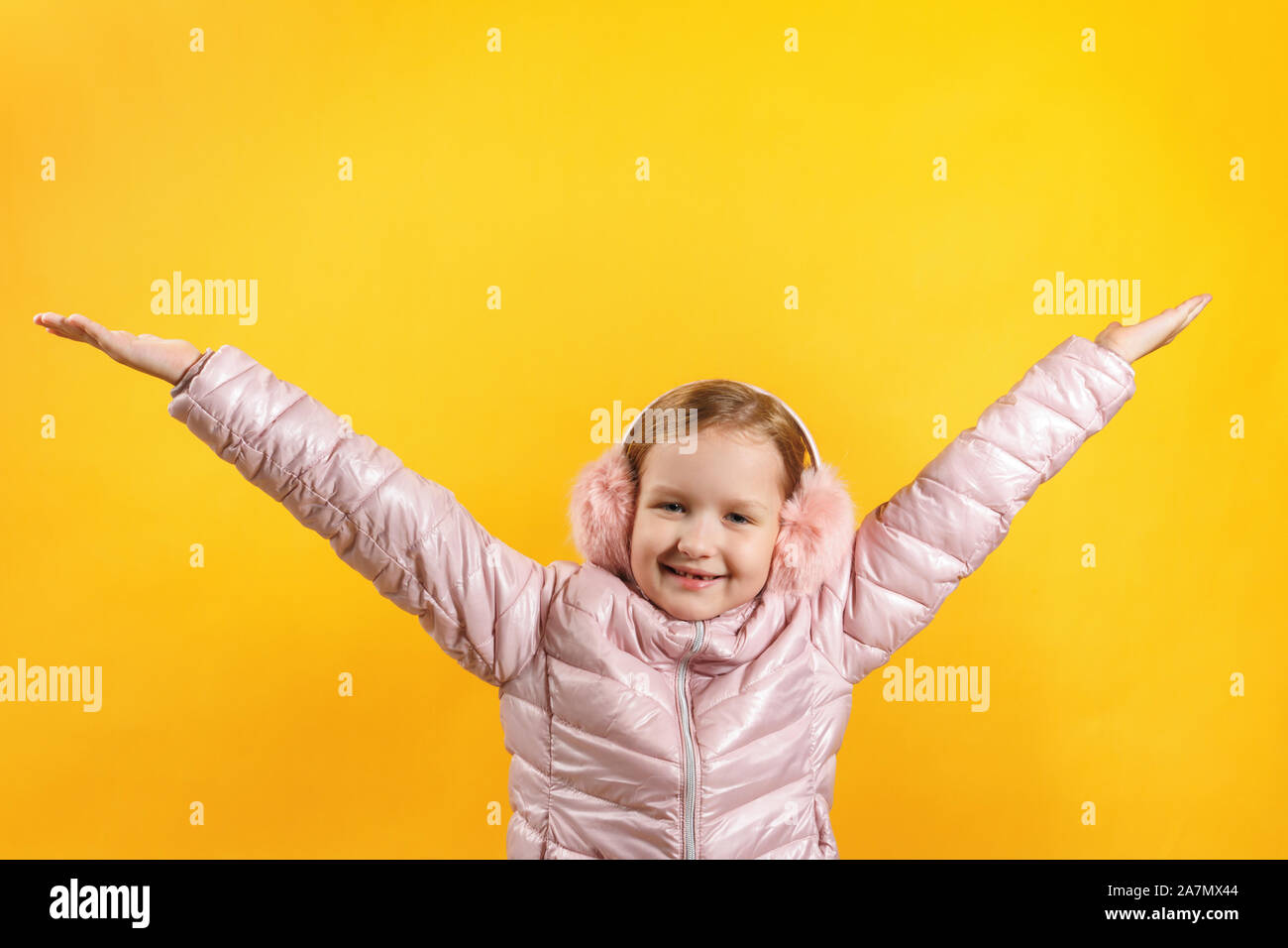 The child in a jacket and warm headphones raised her hands up. Little girl on a yellow background. The concept of happiness, winter, autumn. Stock Photo