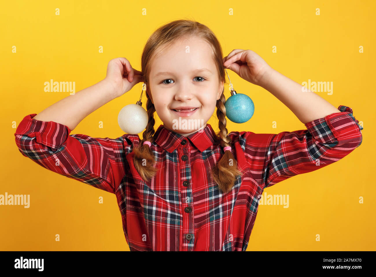 Funny little girl with christmas ball toys near the ears. Close-up of a child in a red dress on a yellow background. Stock Photo