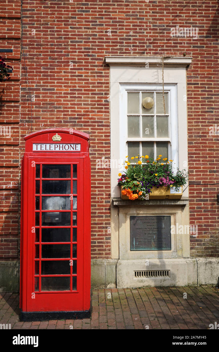 Iconic red British telephone box in front of a brick building with window and flower box on a bright, sunny day, Sidmouth, Devon, UK Stock Photo