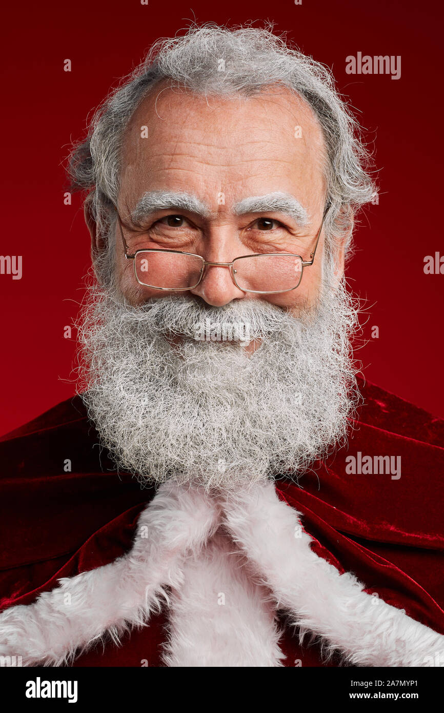 Head and shoulders portrait of classic bearded Santa looking at camera and smiling while posing against red background in studio, copy space Stock Photo