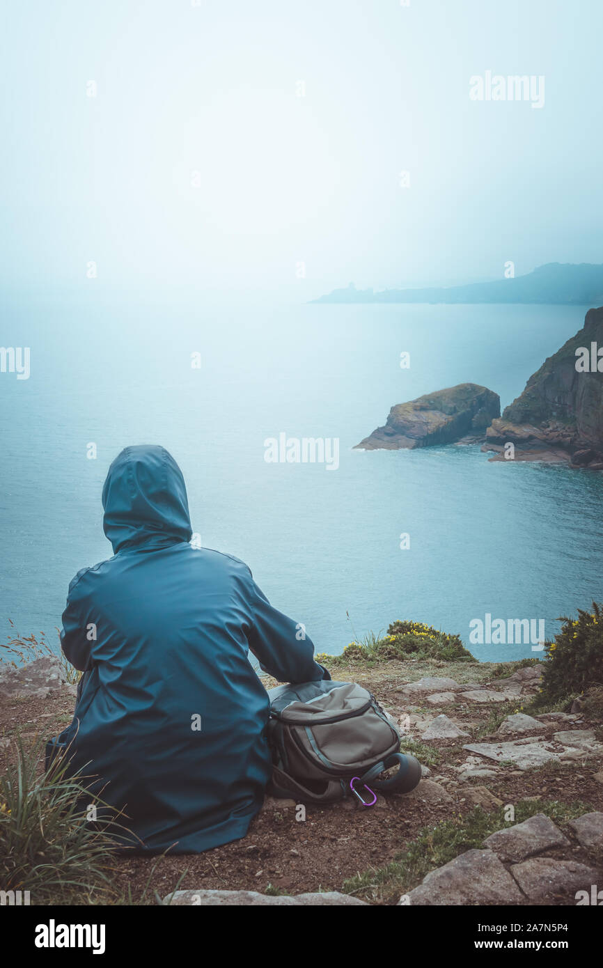 Rear view of hiker woman sitting on the edge of a cliff with blue raincoat and backpack looking at the horizon and sea. Stock Photo