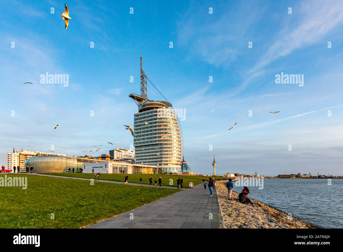 Bremerhaven, Germany - 2 Nov 2019: People playing with seagulls on Weser river Promenade am Strom riverfront embarkment in sunset. Sail-shaped Atlanti Stock Photo