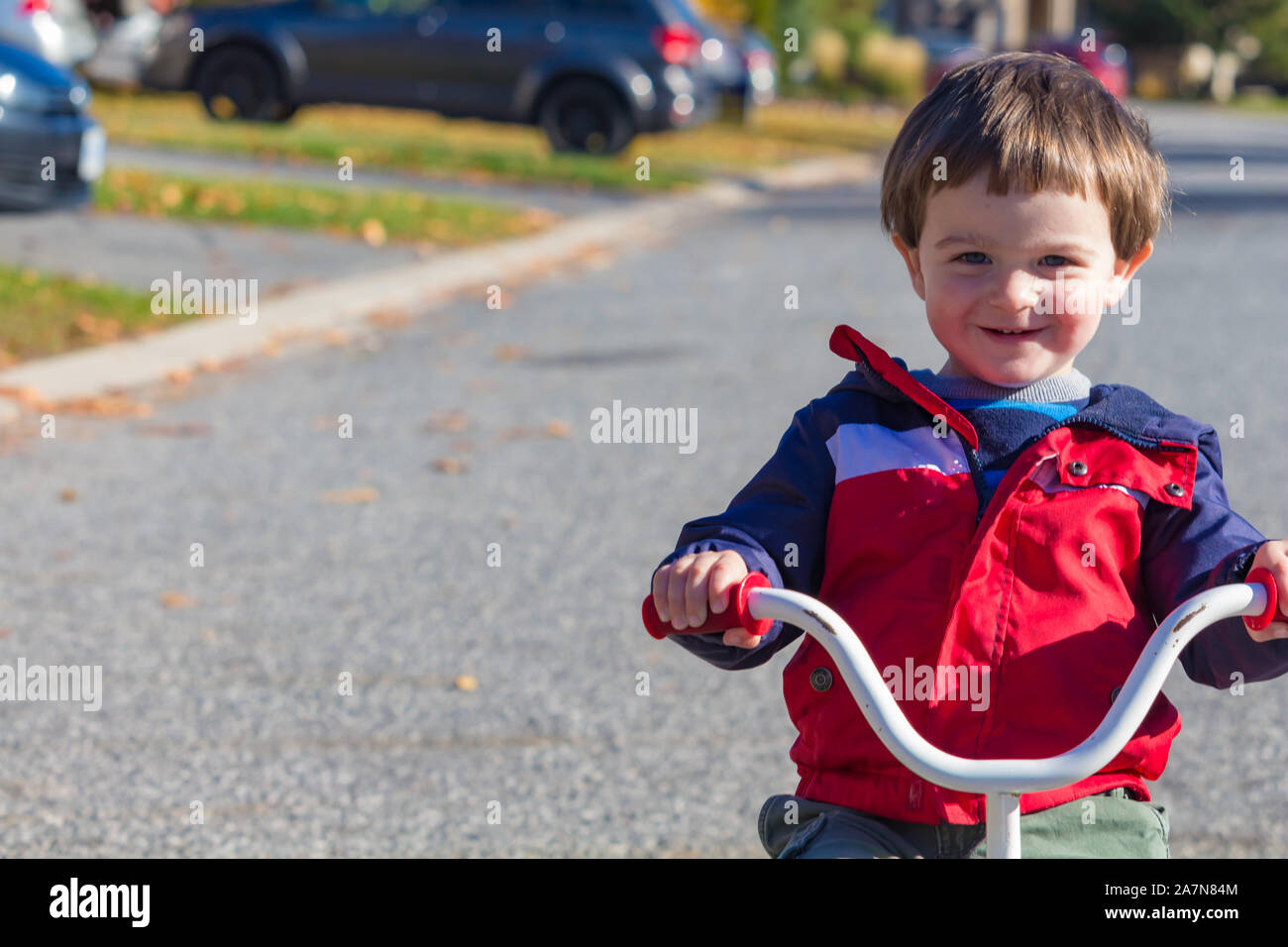 A young toddler, a 2-year-old, rides a tricycle down a suburban street. The little boy smiles as he goes, wearing a light jacket for fall or spring we Stock Photo