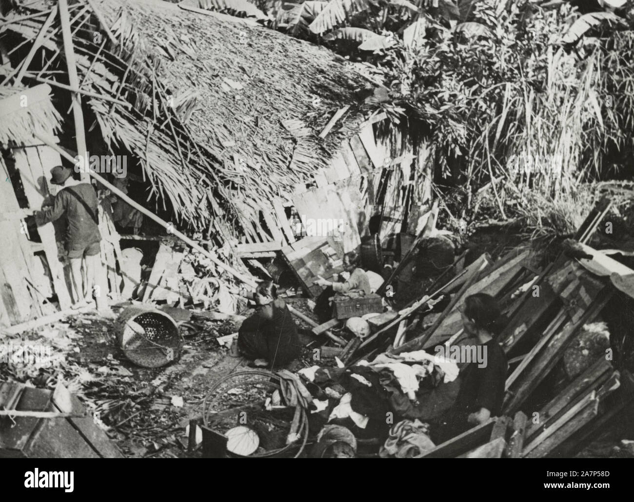 A Malay Chinaman erecting a temporary shelter amid the ruins of his home bombed by Japanese raiders, while his wife (right) collects their scattered possessions. February 1942 Stock Photo