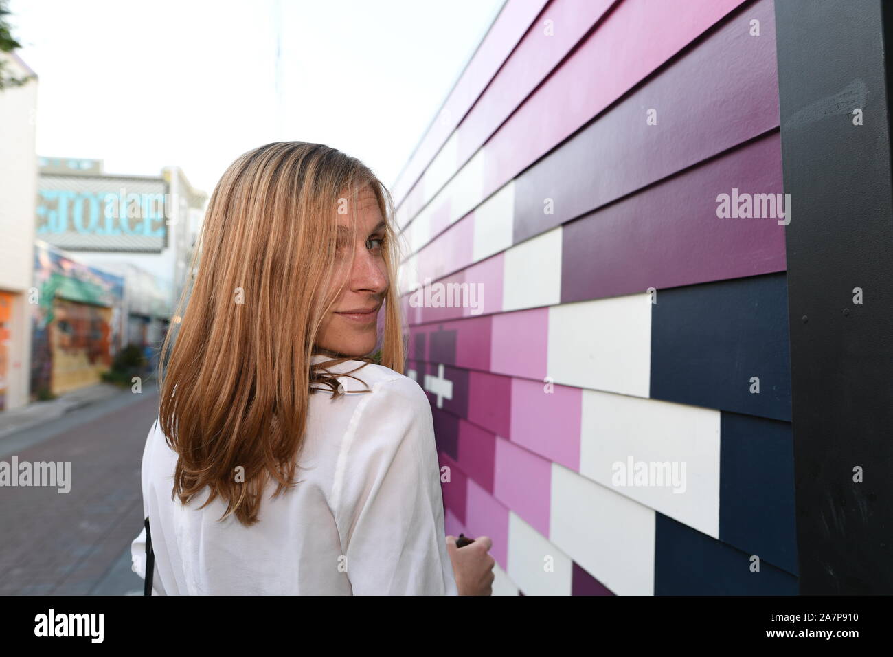 young blonde woman looking over her shoulder wearing a white shirt, walking past large purple painted design on wall siding in an urban alley Stock Photo